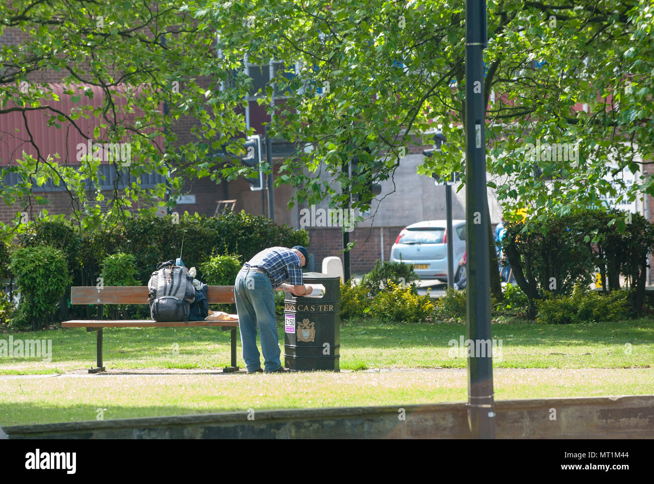 Un uomo perquisisce un bidone in un parco pubblico in una giornata di sole, evidenziando la povertà urbana e i problemi sociali in un ambiente urbano, nel Regno Unito Foto Stock