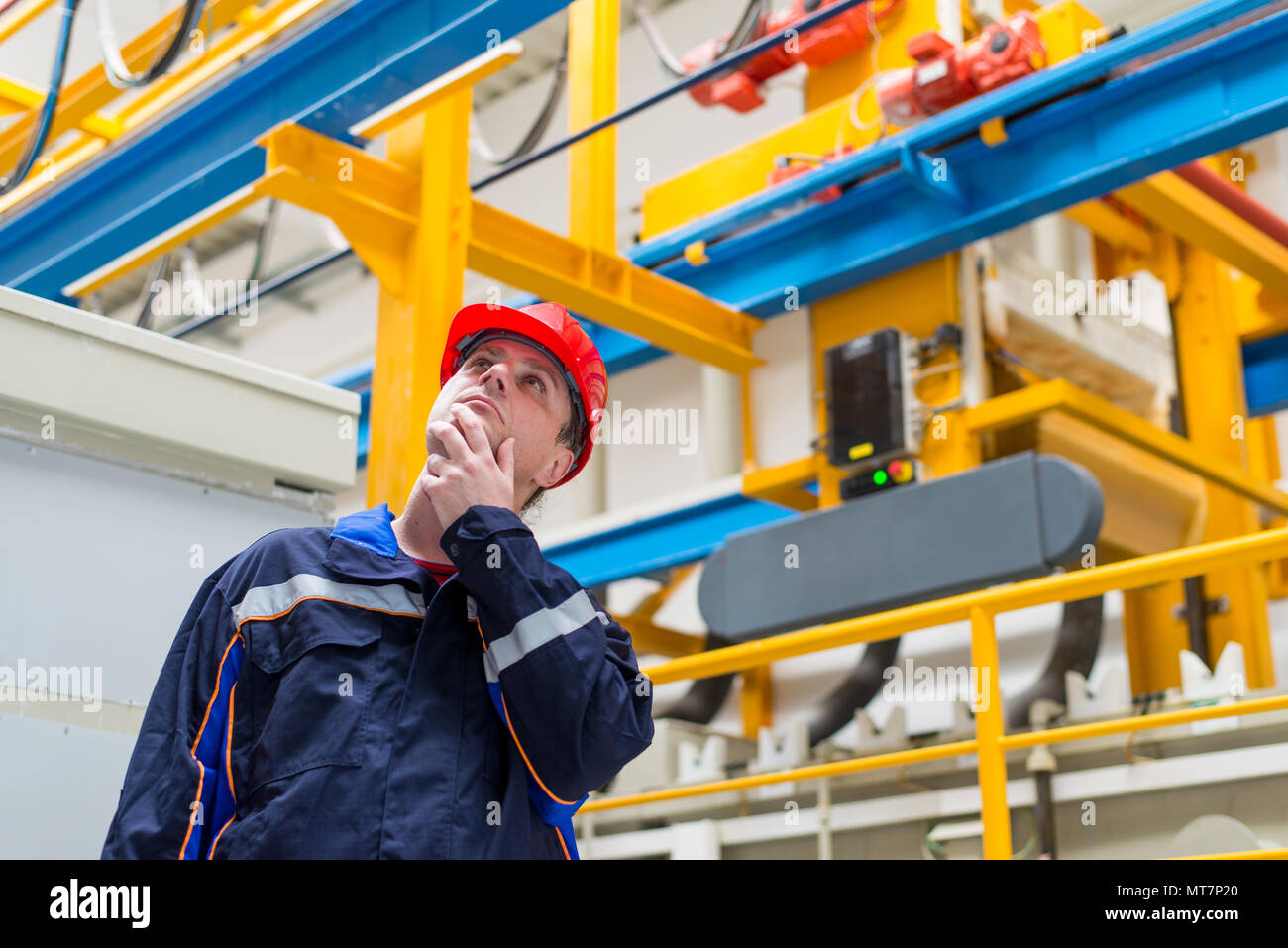 Lavoratore in una fabbrica che indossa blu di lavoro tuta e casco rosso cercando in macchine in background Foto Stock