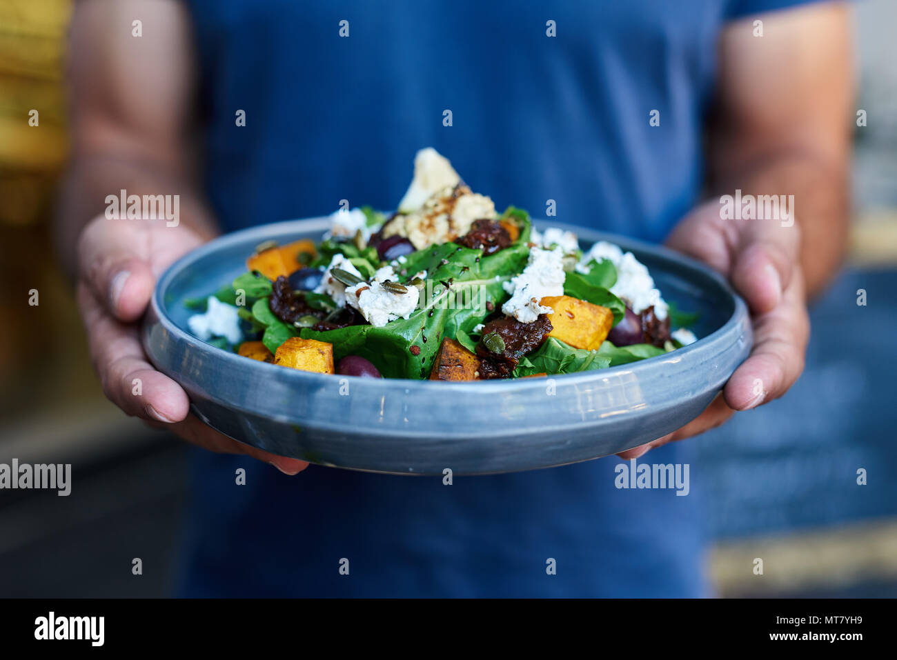 Primo piano di un server in piedi al di fuori di un bistro tenendo una piastra con una deliziosa insalata mista di butternut, lattuga, olive e formaggio feta Foto Stock