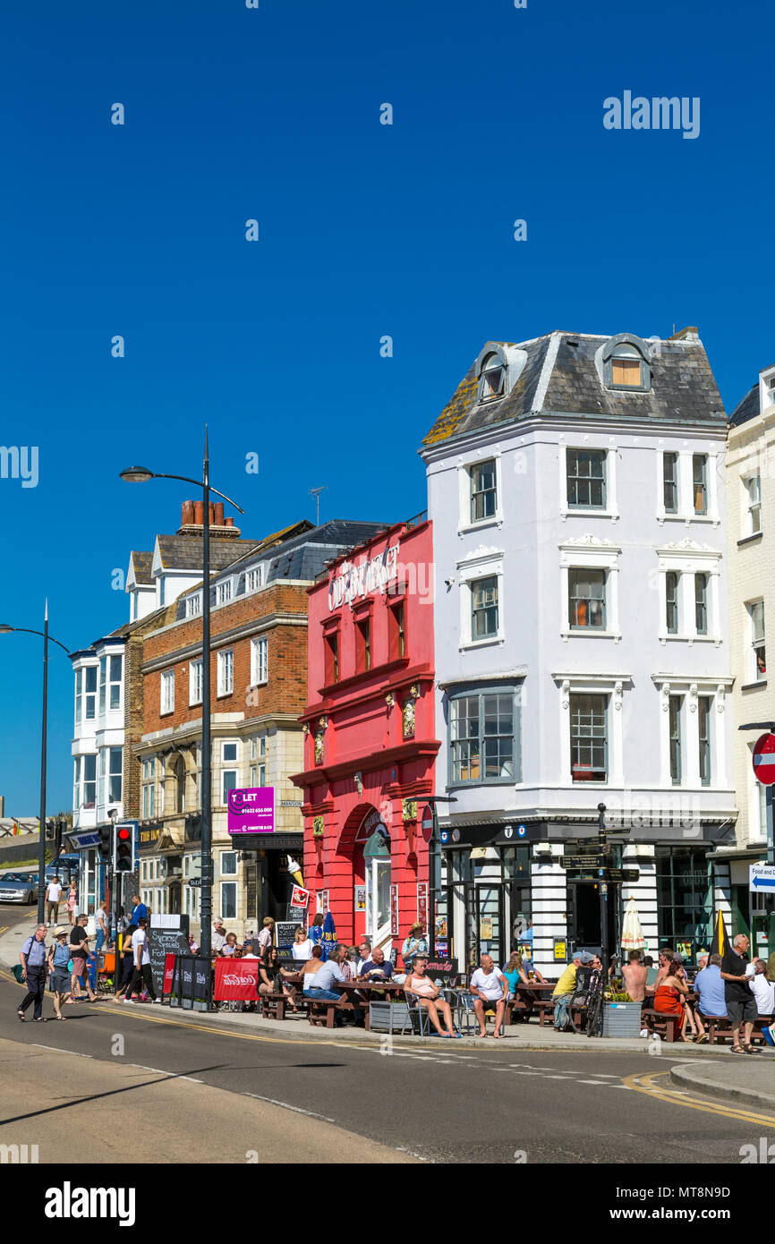 La gente seduta fuori a bere al fresco in un lungomare fronte strada (la parata) case e Old Kent Market Building (rosso) in Margate, Kent, Regno Unito Foto Stock