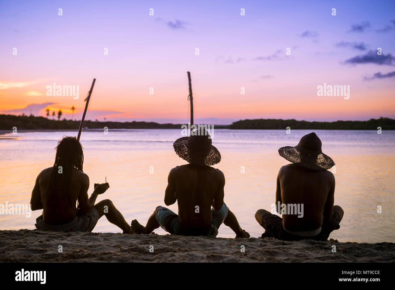 Sagome di tre irriconoscibile capoeiristas sat sulla spiaggia pagando berimbau e percussioni mentre guardate il tramonto a Bahia, Brasile. Foto Stock