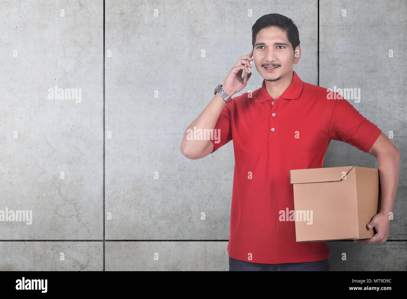 Allegro asiatica corriere maschio utilizzando il cellulare mentre si trattiene il pacco oltre il muro grigio sfondo Foto Stock
