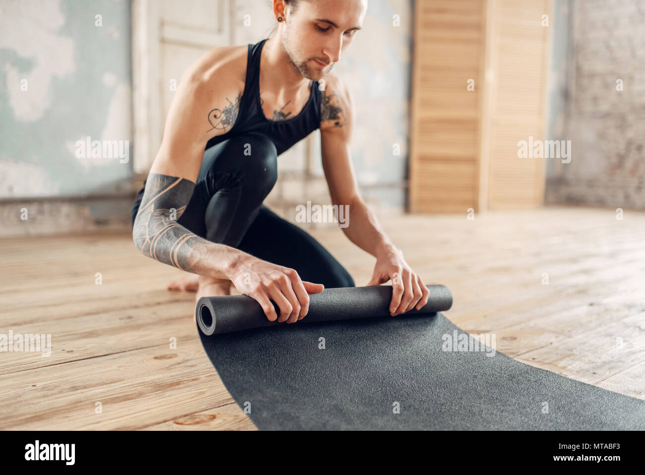 Yoga maschio con tatuaggio sul lato prepara il tappetino per allenamento in palestra con grunge interni. Montare l'allenamento al chiuso Foto Stock