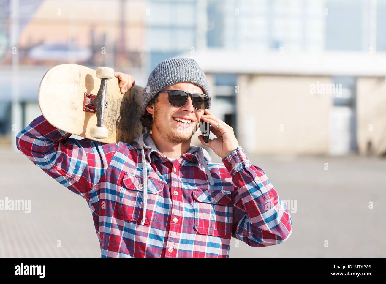 Ritratto di hipster Uomo con occhiali da sole vestiti in maglietta cheked e trattenimento tappo skateboard sulla spalla e parlando da smartphone prima di edificio moderno Foto Stock