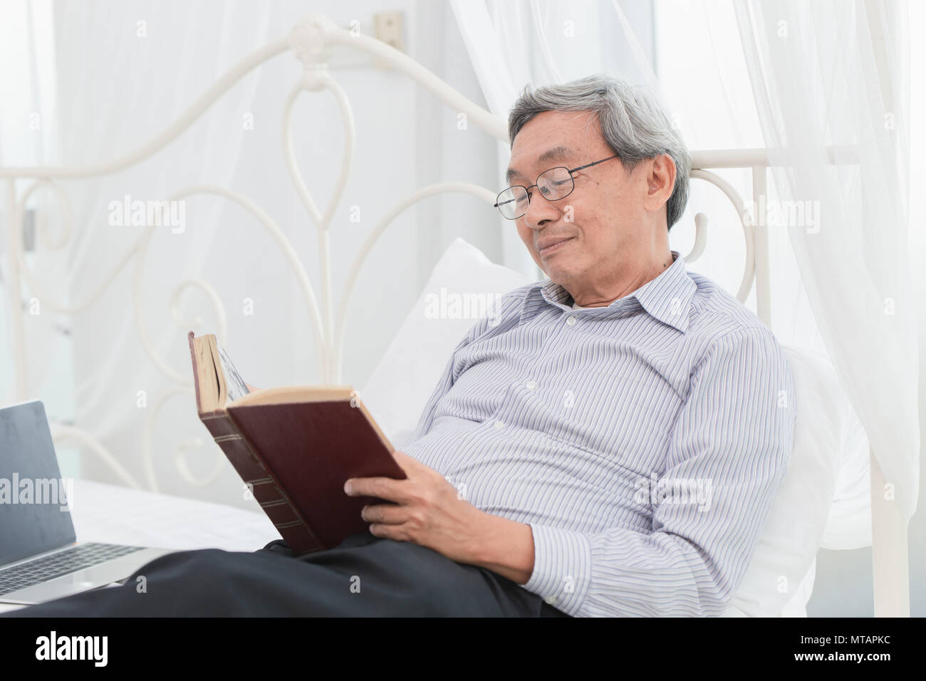 Asian vecchio professore occhiali godetevi la lettura di libro di testo ogni mattina sul letto Foto Stock