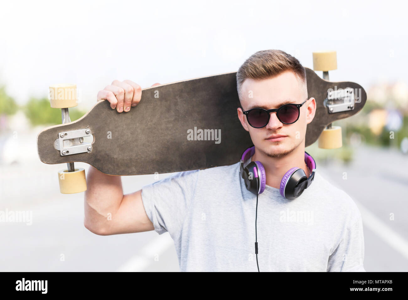 Guidatore di skateboard vestito in grigio di t-shirt e occhiali da sole con auricolari detiene longboard sulla spalla su una strada di città Foto Stock