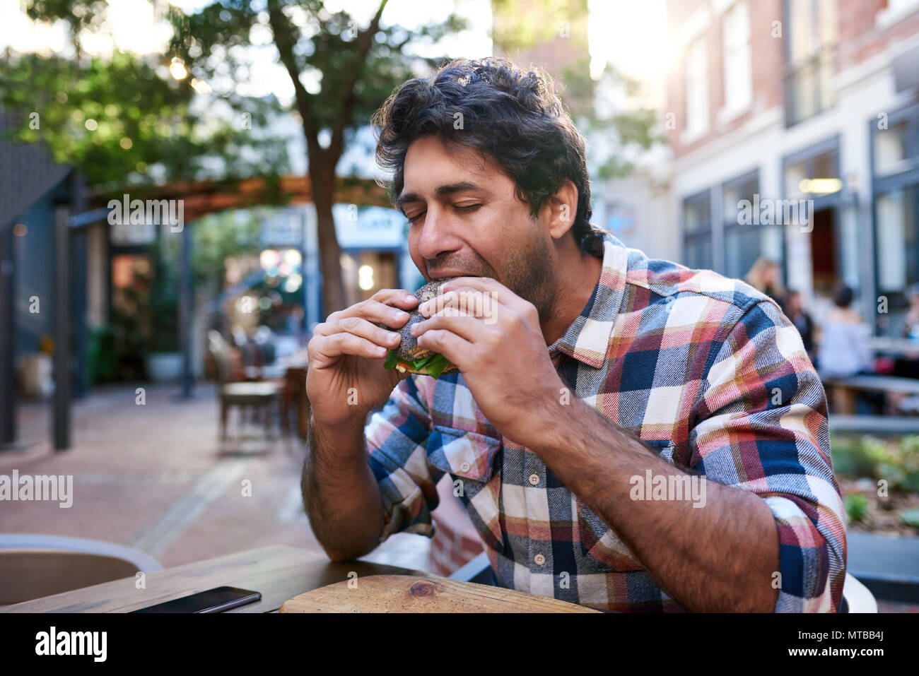 Fame giovane uomo prendendo un morso del suo seme di papavero bagel seduti con gli occhi chiusi al di fuori di un cafè sul marciapiede tabella Foto Stock