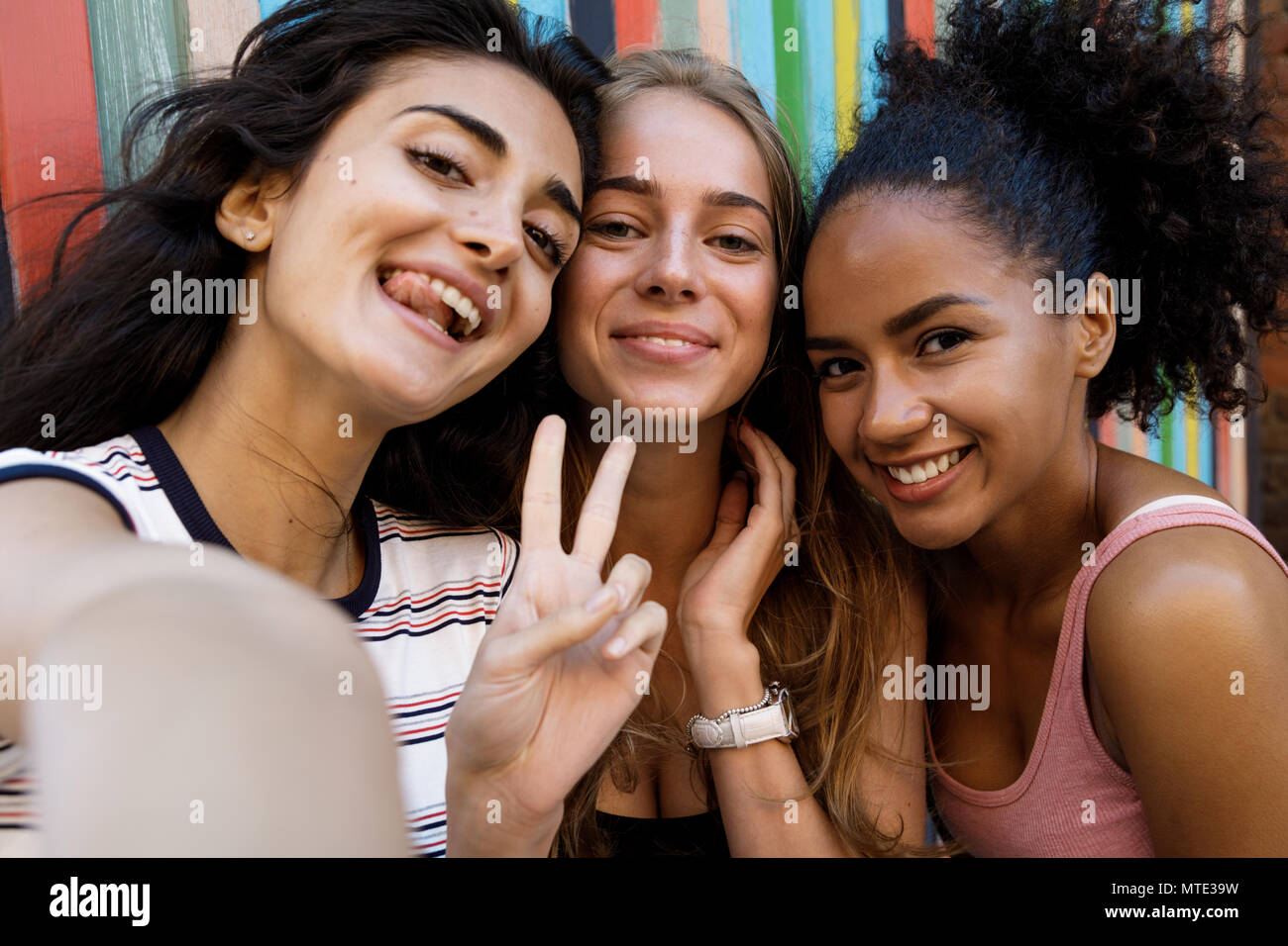Tre felici donne rendendo selfie, in piedi su una strada di città a parete colorata Foto Stock