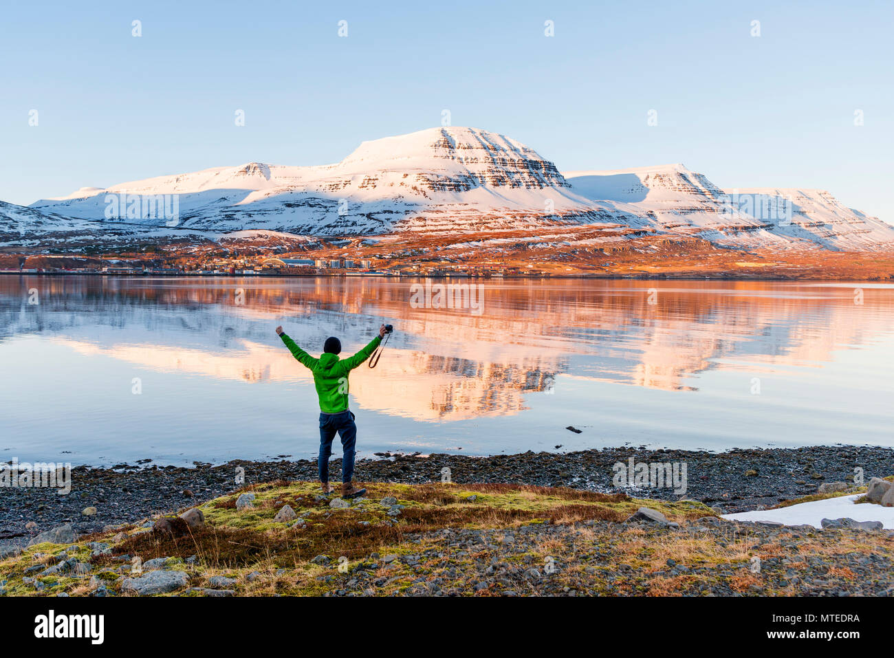 Uomo in piedi sulla riva stretching le braccia in aria, acqua riflessione, montagne innevate in un fiordo, Reyðarfjörður Foto Stock