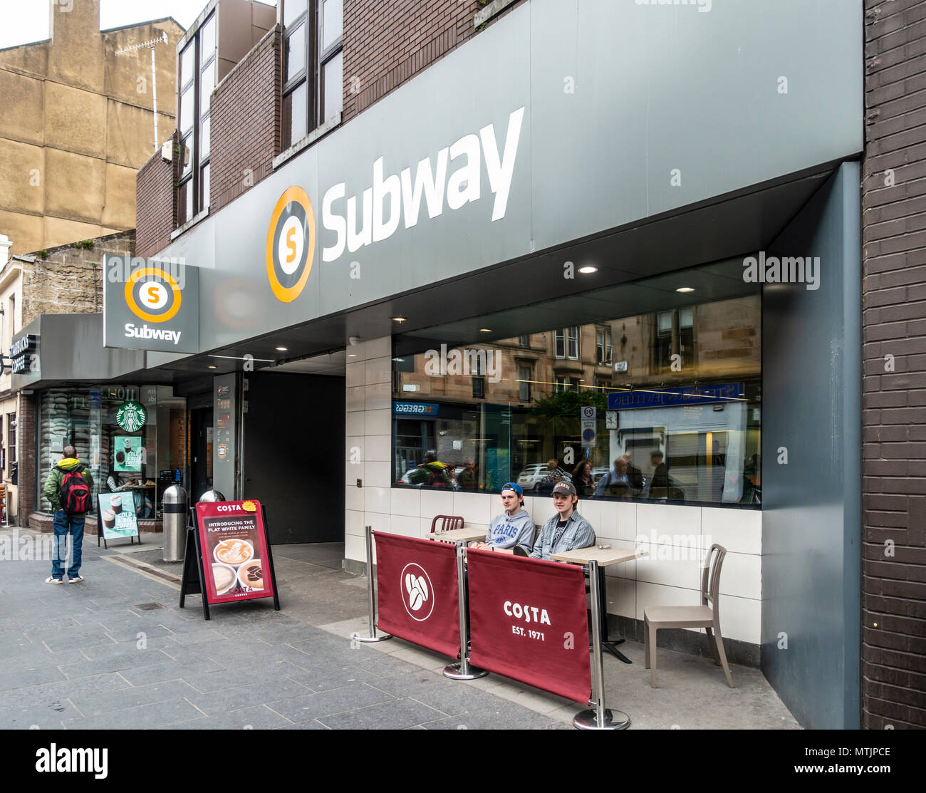 Due giovani uomini in Costa all'esterno area con posti a sedere di fronte Hillhead Stazione della Metropolitana in Byres Road, Glasgow; un altro uomo guarda attraverso la Starbuck Foto Stock