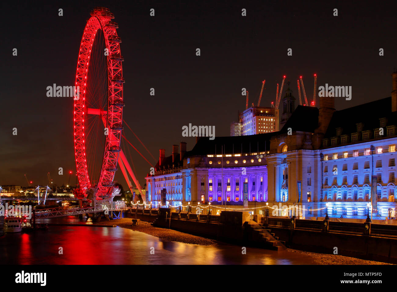 Il London Eye, County Hall e del Tamigi illuminato con il rosso e il blu di illuminazione, più gru dominano lo skyline dove più lavori di costruzione in corso Foto Stock