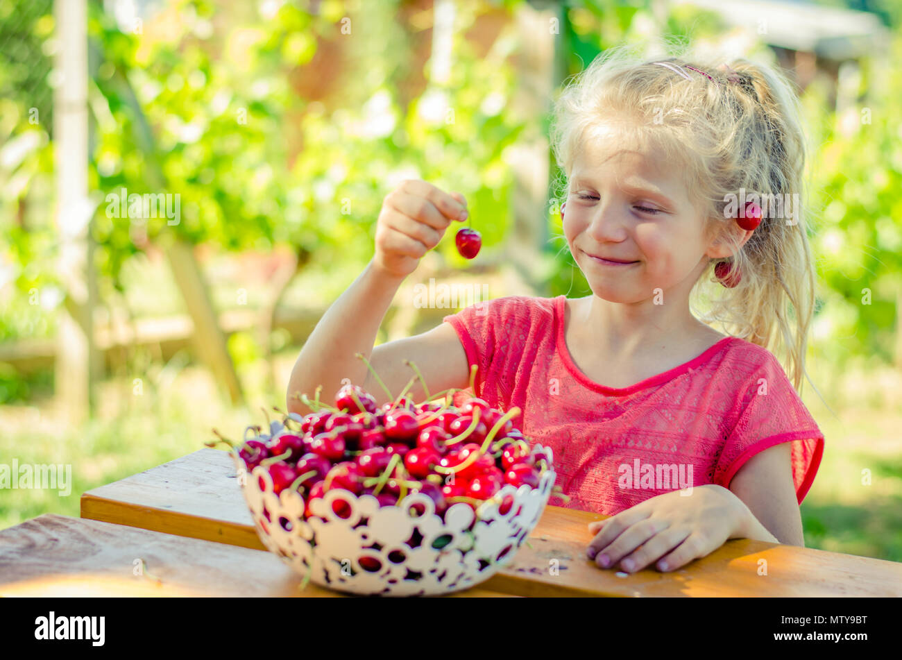 Adorabile ragazza bionda mangiare frutta di ciliegio in giardino Foto Stock