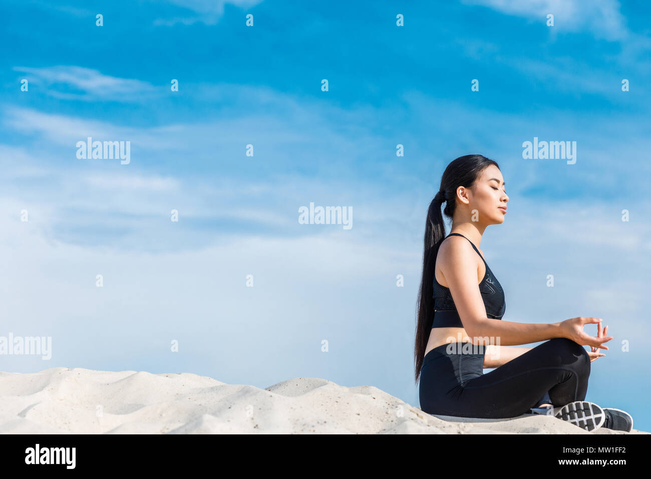 Donna asiatica meditando in lotus yoga pone con gyan mudra sulla sabbia contro il cielo nuvoloso Foto Stock