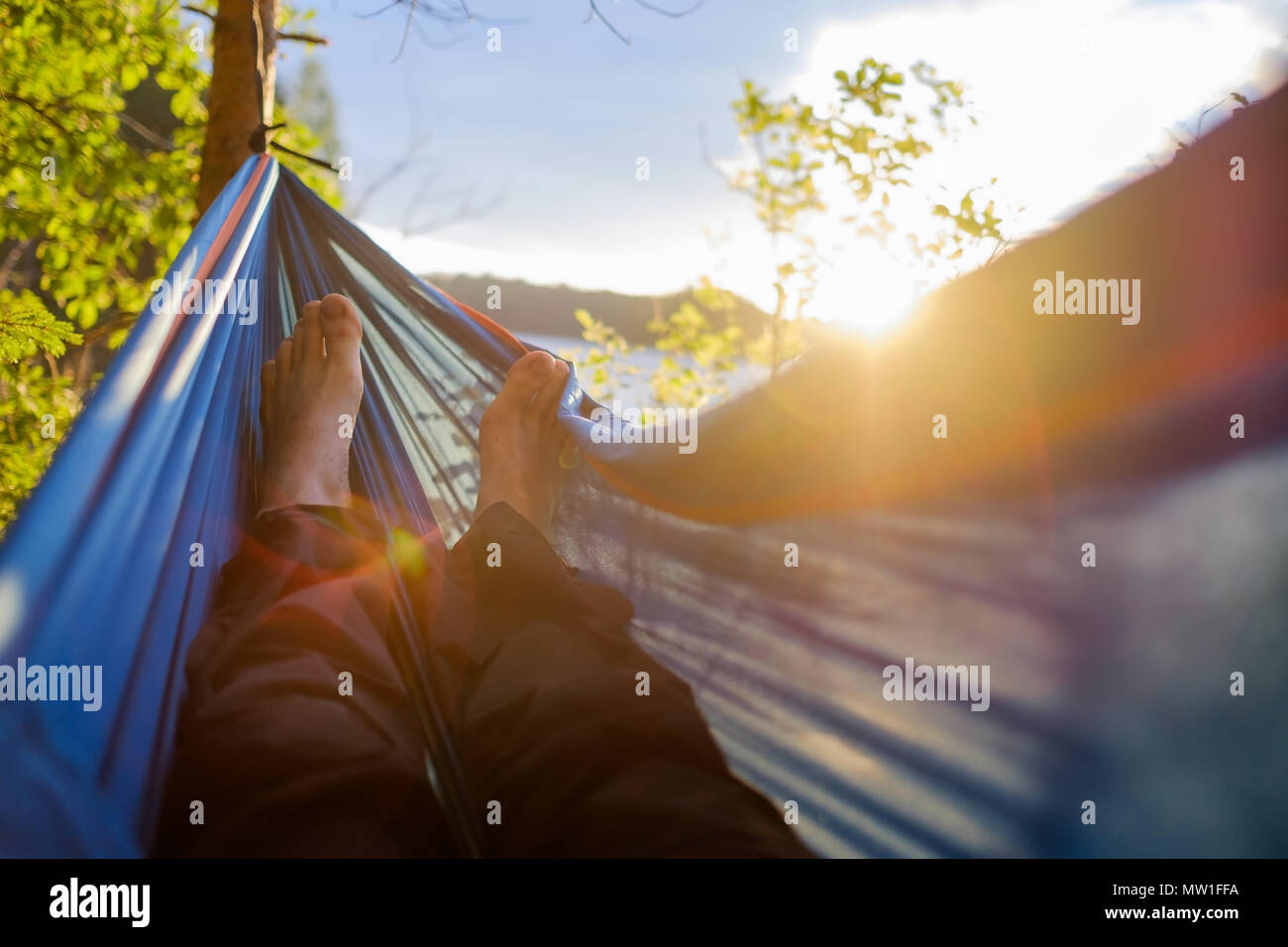 L'uomo i piedi sono visti da vicino da un amaca in una Foresta estate, su uno sfondo sfocato di pini e di un lago. Foto Stock