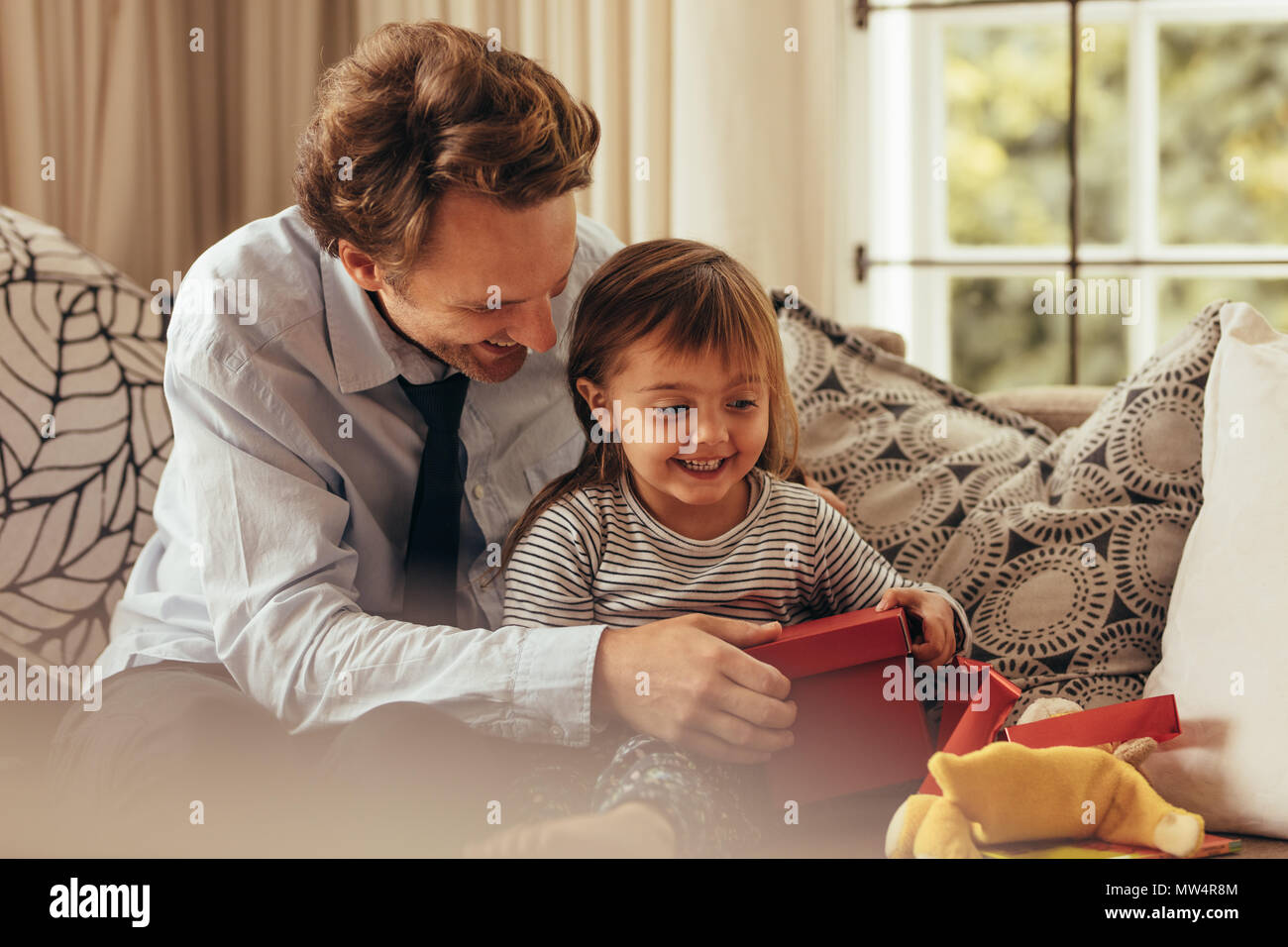 Padre e figlia seduta su un divano apertura di una confezione regalo. Aiutando il padre di sua figlia apertura di una confezione regalo. Foto Stock
