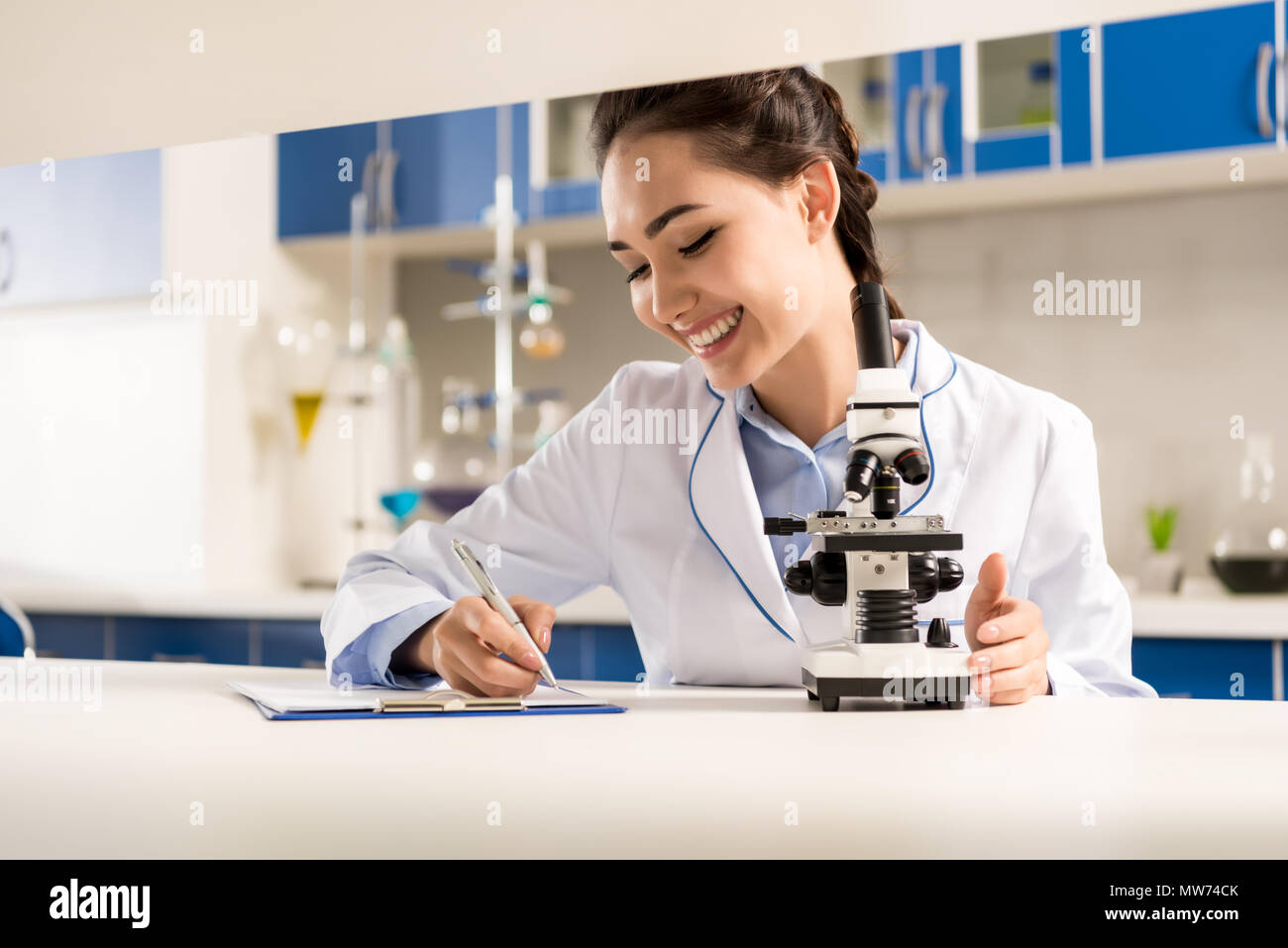 Giovani sorridente tecnico di laboratorio prendendo appunti dopo il microscopio facendo analisi del campione Foto Stock