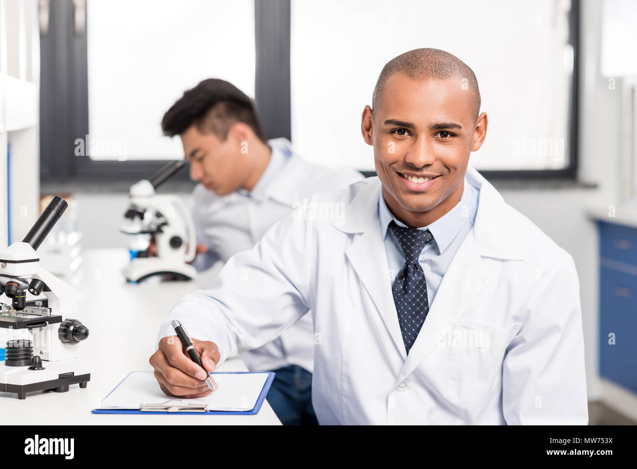 Bel giovane medico in camice scrivere appunti su appunti e sorridente Foto Stock