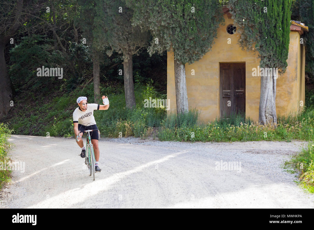 Ciclista in bicicletta passando accanto alla chiesetta e ai cipressi di Lucignano d'Asso, partecipando a maggio all'Eroica Montalcino, Siena, Toscana, Italia Foto Stock