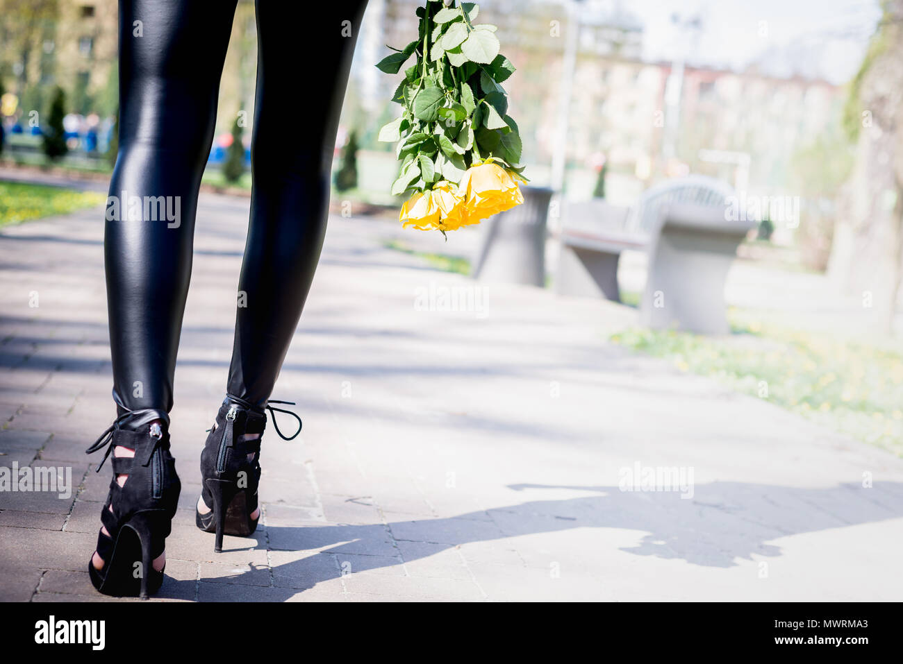 Vista delle gambe di una donna che cammina con fiori. Donna indossare tacchi alti scarpe. La ragazza in pantaloni neri si muove intorno al di fuori del parco. Beuty e concetto di moda. Foto Stock