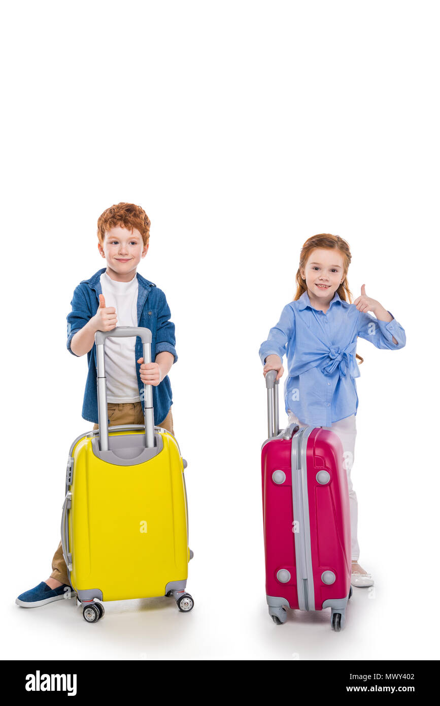 Adorabili e sorridente redhead kids in piedi con le valigie e mostrando pollice in alto isolato su bianco Foto Stock