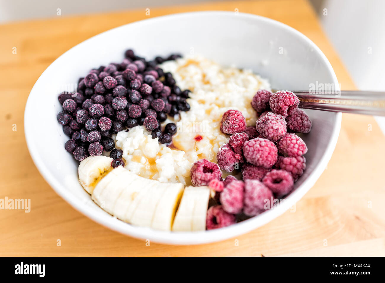 Macro closeup di fiocchi d'avena Grani di Avena budino di riso ciotola di porridge con frutti di bosco surgelati, Lamponi Mirtilli, banane e sciroppo d'acero caramello su legno Foto Stock