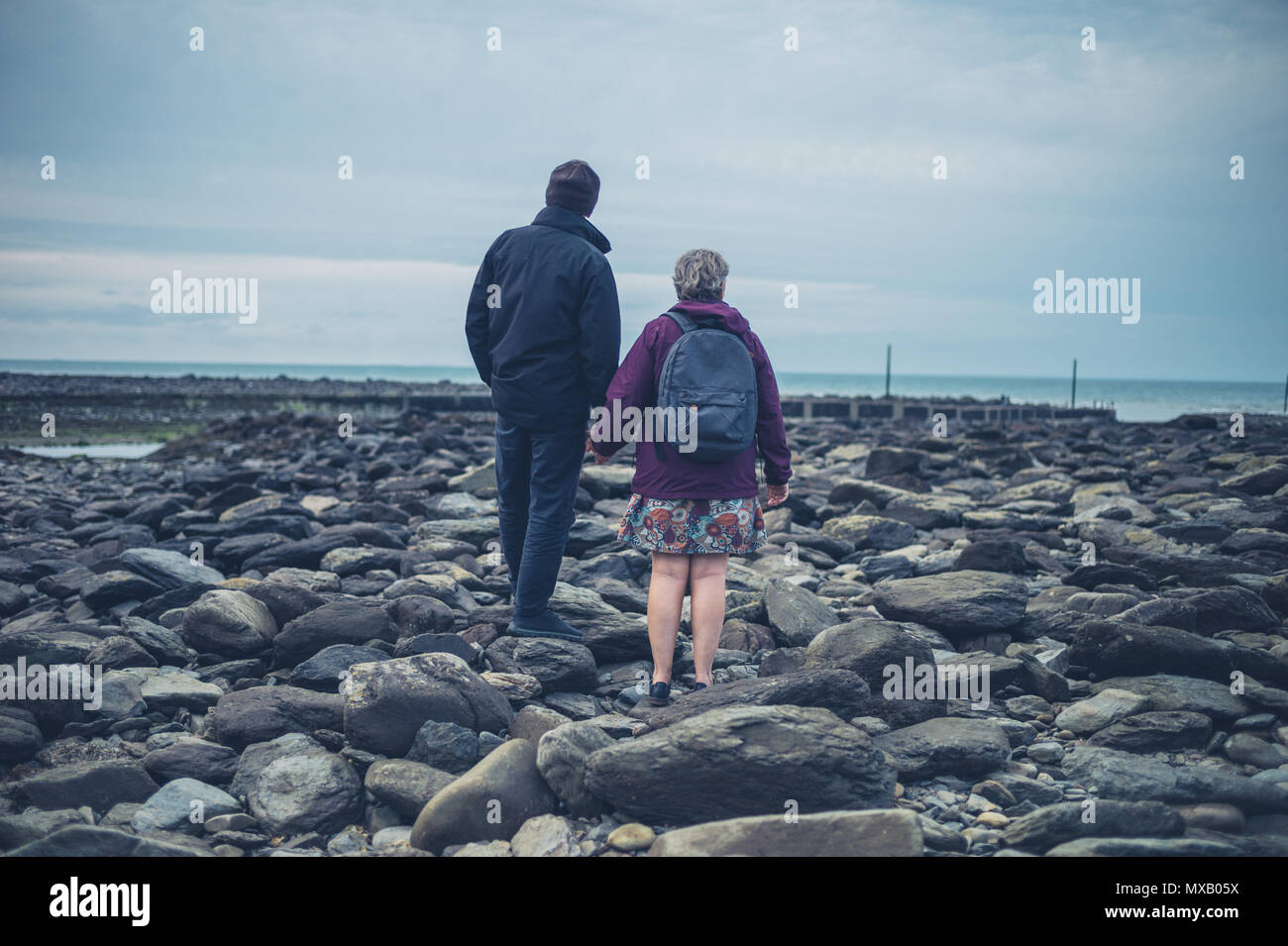 Una coppia senior è in piedi e tenendo le mani su una spiaggia rocciosa in autunno Foto Stock