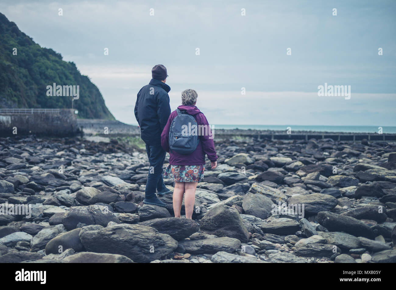 Una coppia senior è in piedi e tenendo le mani su una spiaggia rocciosa in autunno Foto Stock