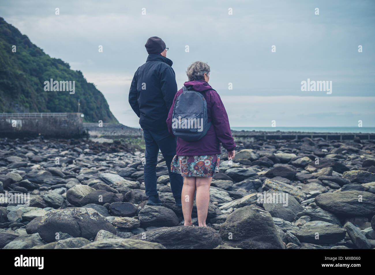 Una coppia senior è in piedi e tenendo le mani su una spiaggia rocciosa in autunno Foto Stock