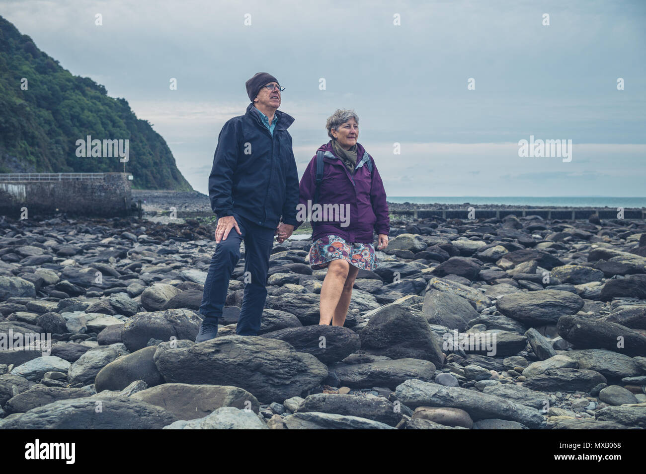 Una coppia senior è in piedi e tenendo le mani su una spiaggia rocciosa in autunno Foto Stock