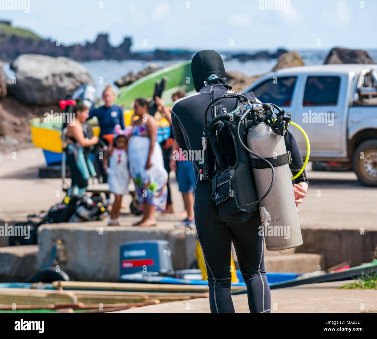 Uomo in portante muta scuba serbatoio di ossigeno e per le immersioni a piedi barca in porto con la popolazione locale sulla banchina, Hanga Roa, Isola di Pasqua, Cile Foto Stock