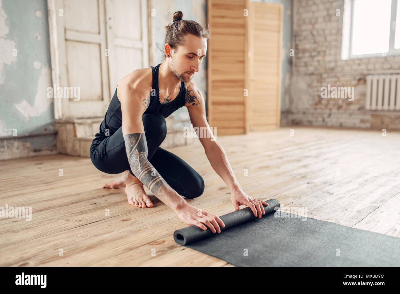 Yoga maschio con tatuaggio sul lato prepara il tappetino per allenamento in palestra con grunge interni. Montare l'allenamento al chiuso Foto Stock