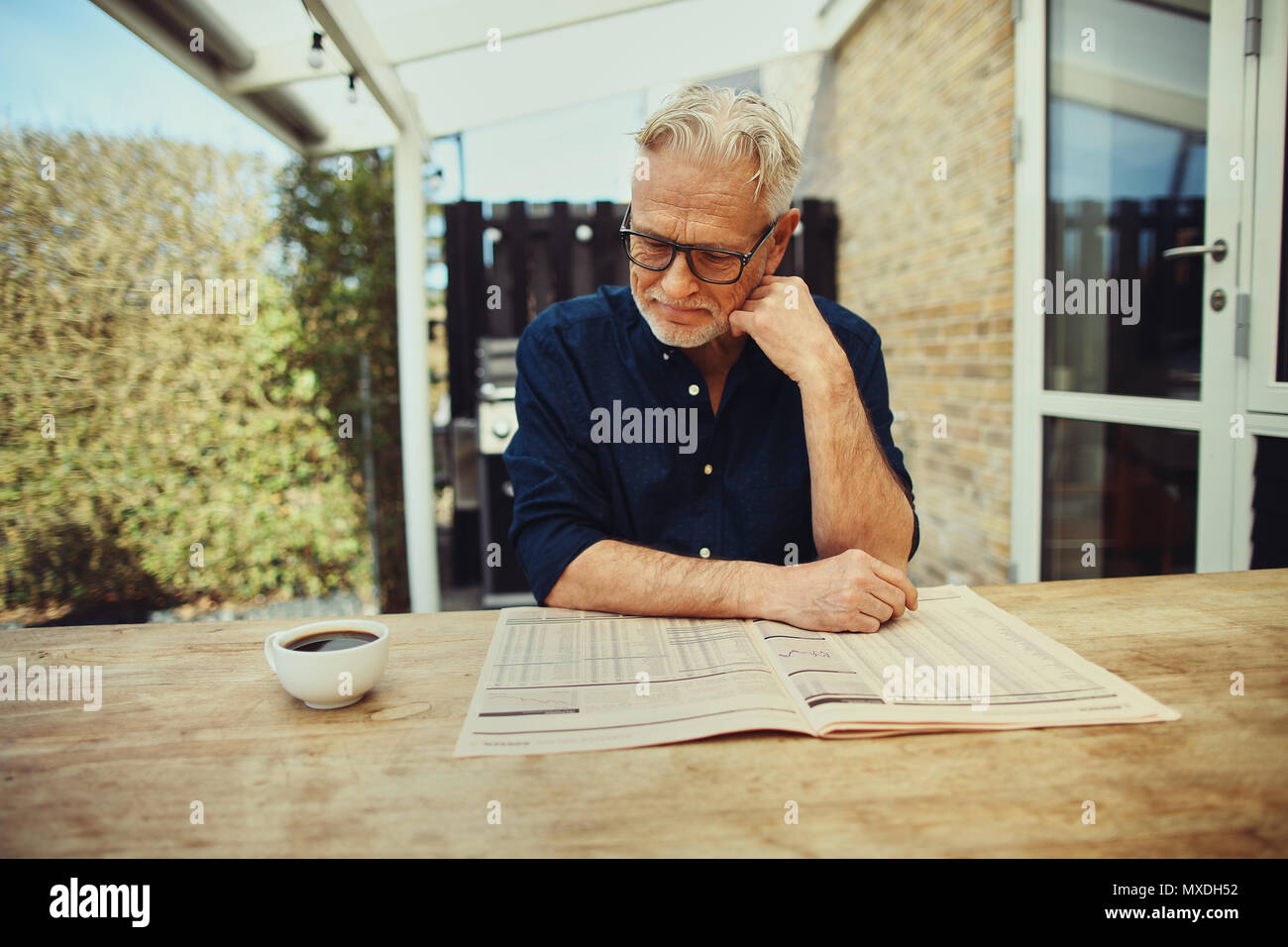 Sorridente uomo senior leggendo le pagine finanziarie in un giornale e bere una tazza di caffè mentre seduto al tavolo del patio esterno Foto Stock