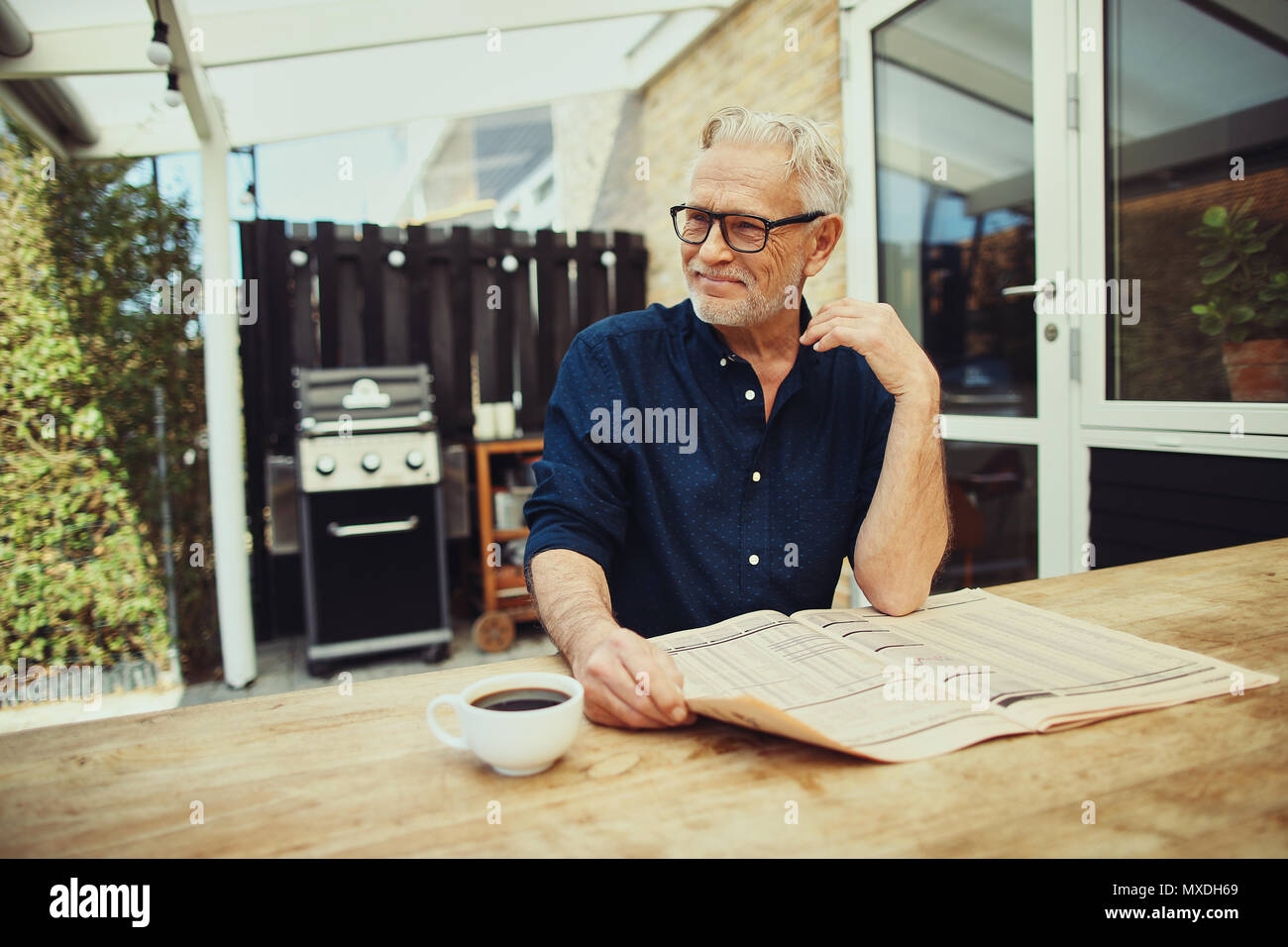 Senior uomo sorridente contently mentre seduto al tavolo del patio di bere una tazza di caffè e la lettura dei giornali Foto Stock