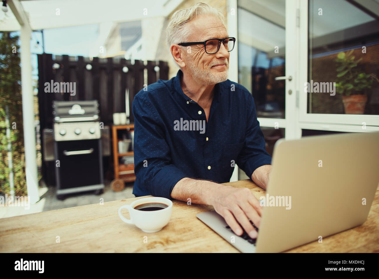 Senior sorridente uomo seduto al tavolo del patio esterno lavorando su un computer portatile e di bere una tazza di caffè Foto Stock