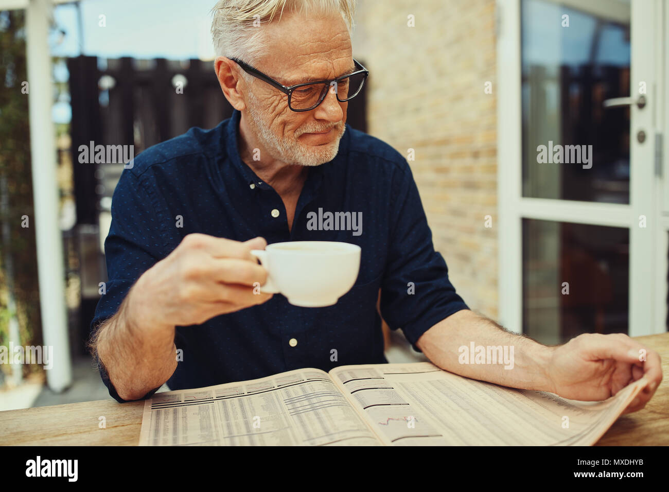 Senior sorridente uomo seduto fuori a tavola sul suo patio leggendo le pagine finanziarie in un giornale e godersi un caffè Foto Stock