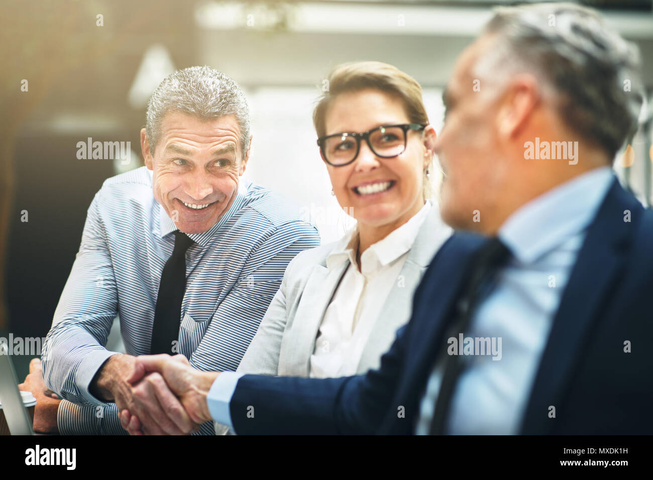 Due imprenditori matura sorridente e agitando le mani insieme con una femmina di un collega a guardare mentre seduti a un tavolo in un ufficio Foto Stock