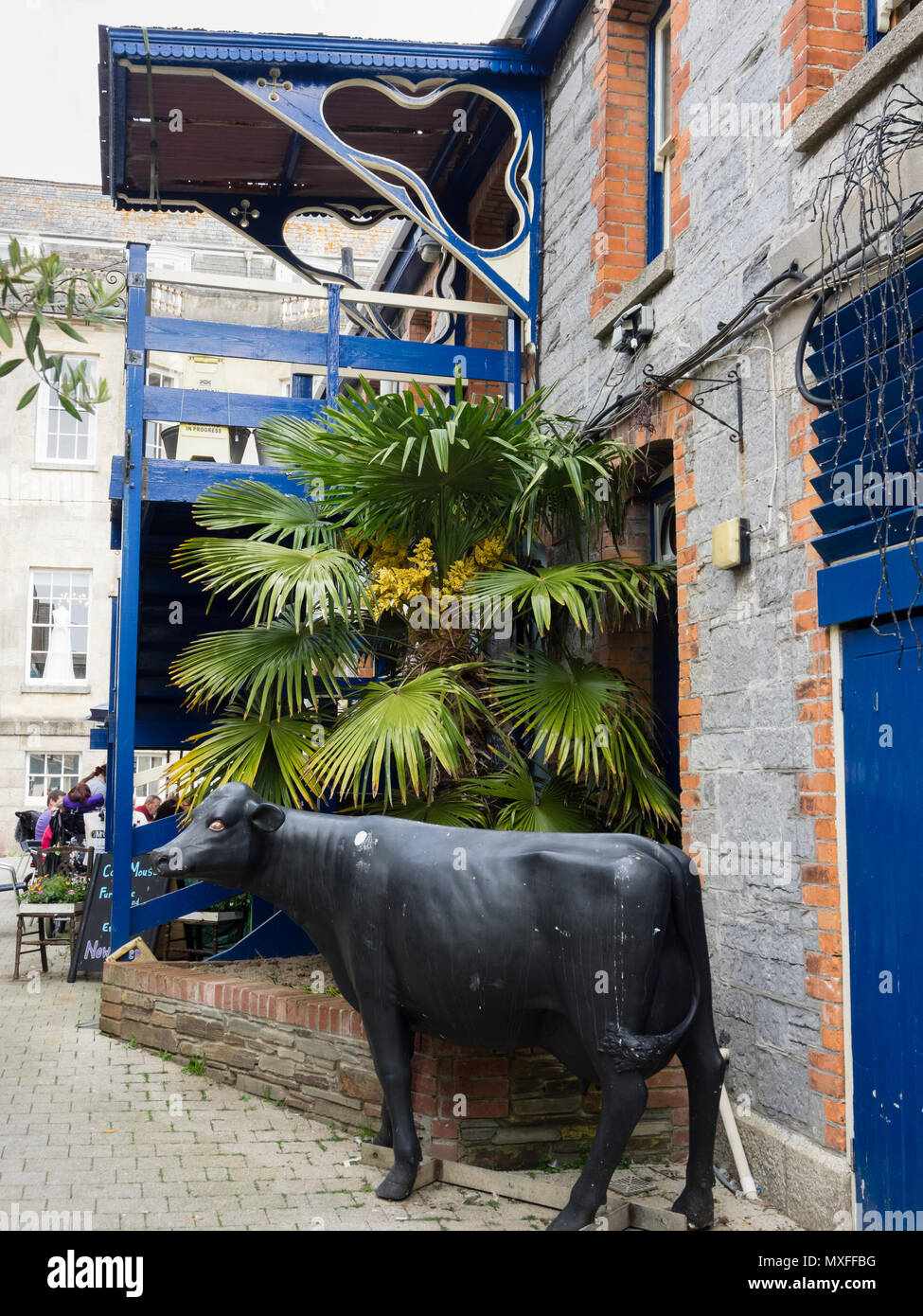 La statua di una mucca nera si erge al di fuori di Truro bauletto di mercato con un balcone blu e trachycarpus fortunei palm dietro Foto Stock