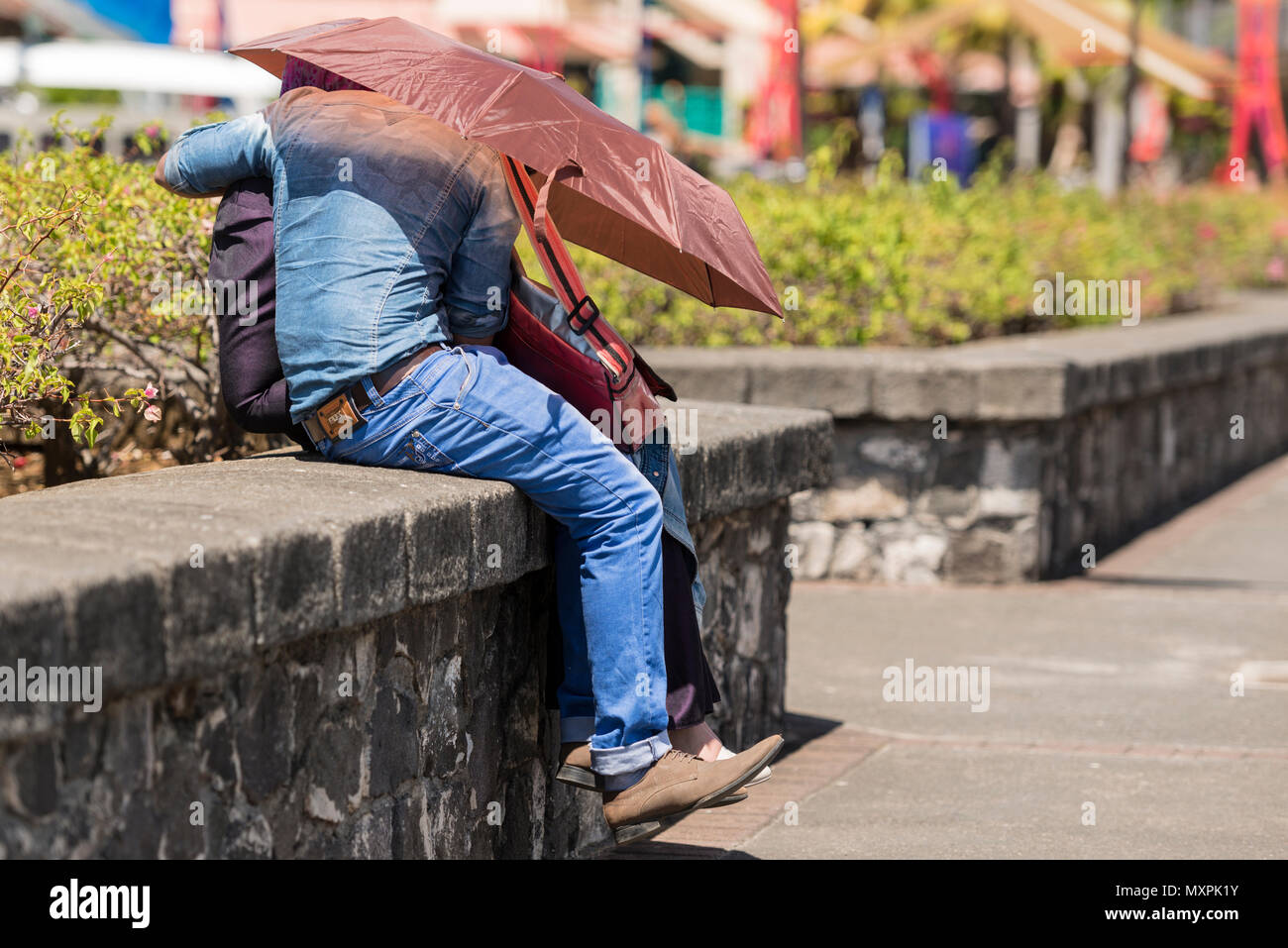 Una giovane coppia discretamente coccola sotto un ombrello sotto il Caudan Waterfront, Port Louis, Maurizio Foto Stock