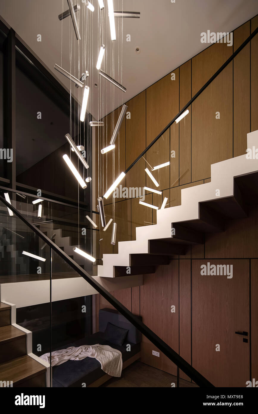 Elegante arredamento moderno con pareti in legno e una scala a pioli in legno e una ringhiera in vetro. Vi sono appesi luminose lampade di fantasia e un divano scuro wi Foto Stock