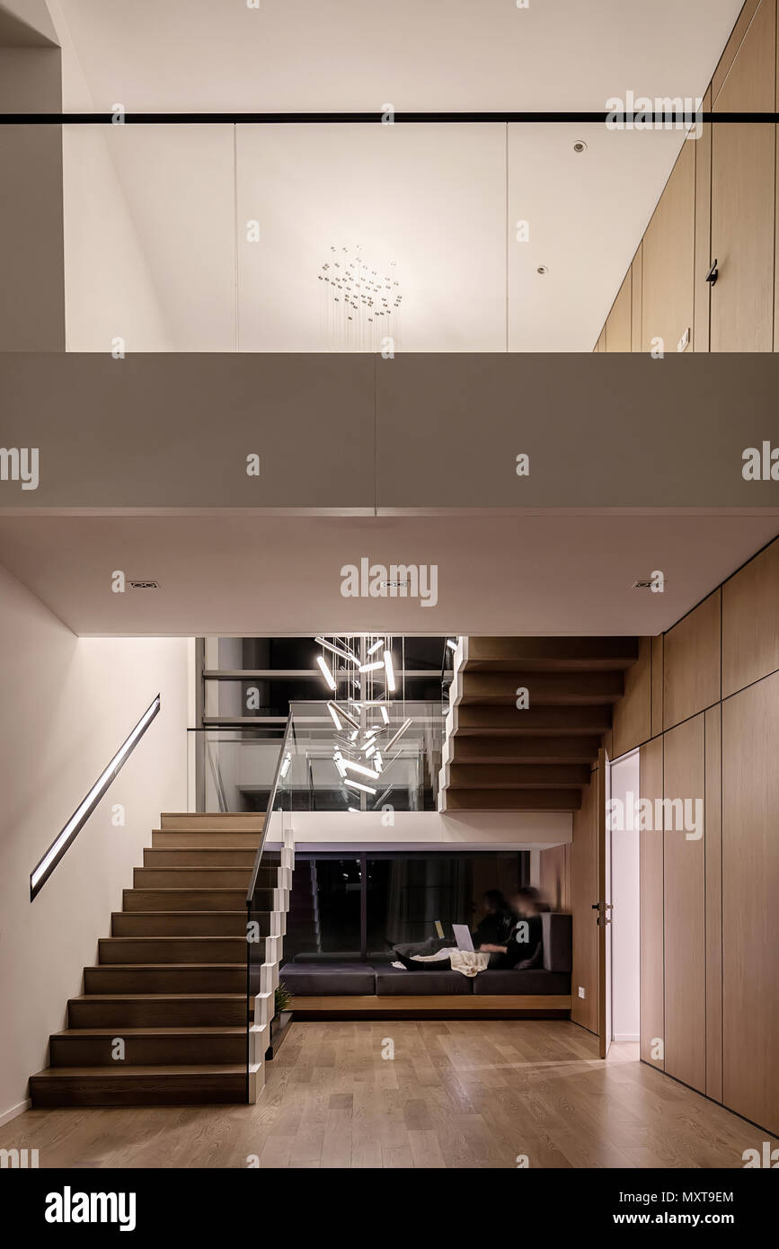 Cool arredamento moderno con pareti chiare e una scala a pioli in legno e una ringhiera in vetro. Vi sono appesi luminose lampade di fantasia e un divano scuro con un Foto Stock
