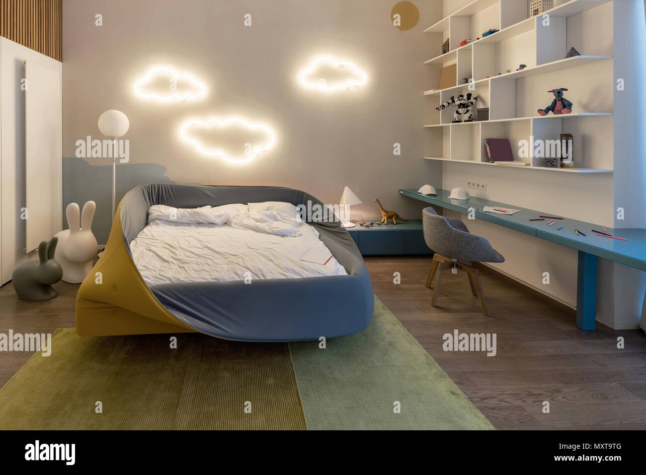 Moderna stanza dei ragazzi con fantasia luminose lampade cloud su una parete bianca. Vi è colorato letto con biancheria bianca, blu tavolo con sedia grigio, bianco shelv Foto Stock