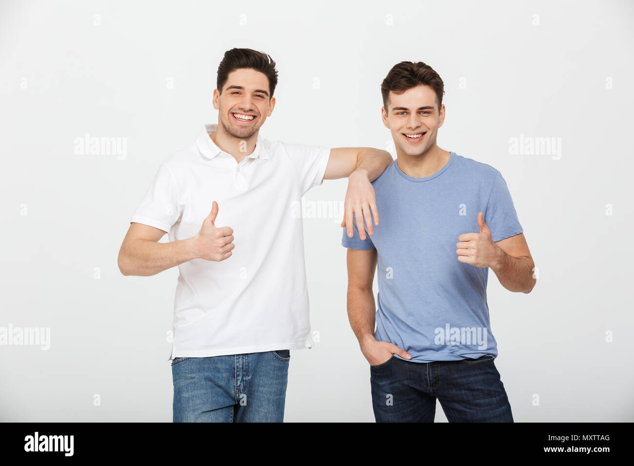 Due amici felice 30s indossando t-shirt casual e jeans sorridente e mostrando pollice in alto sulla fotocamera in studio isolate su sfondo bianco Foto Stock