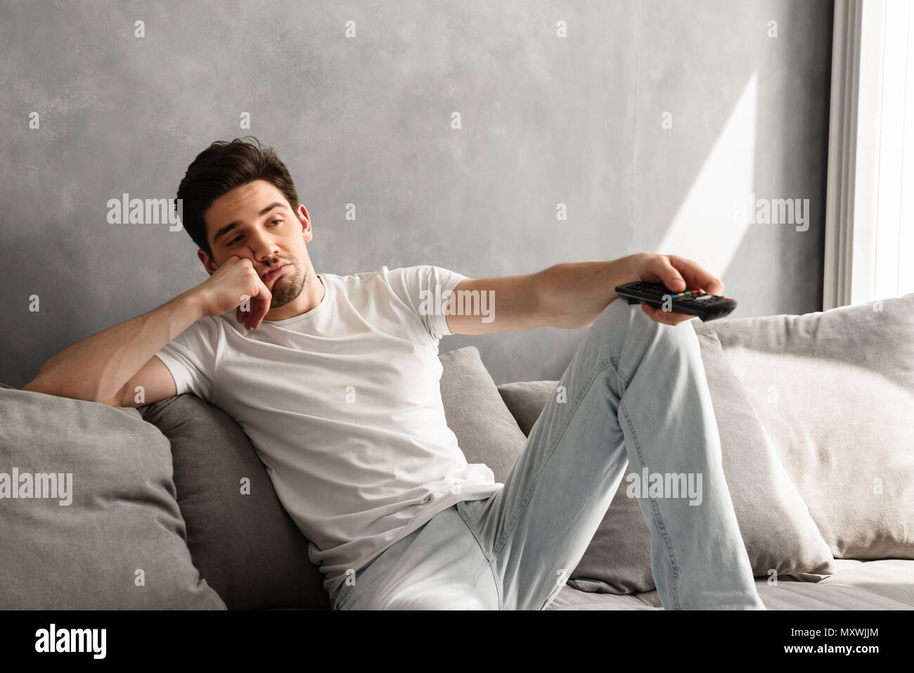 Foto di giovani annoiati uomo seduto sul divano di casa e cambiare i canali televisivi con disinteresse Foto Stock