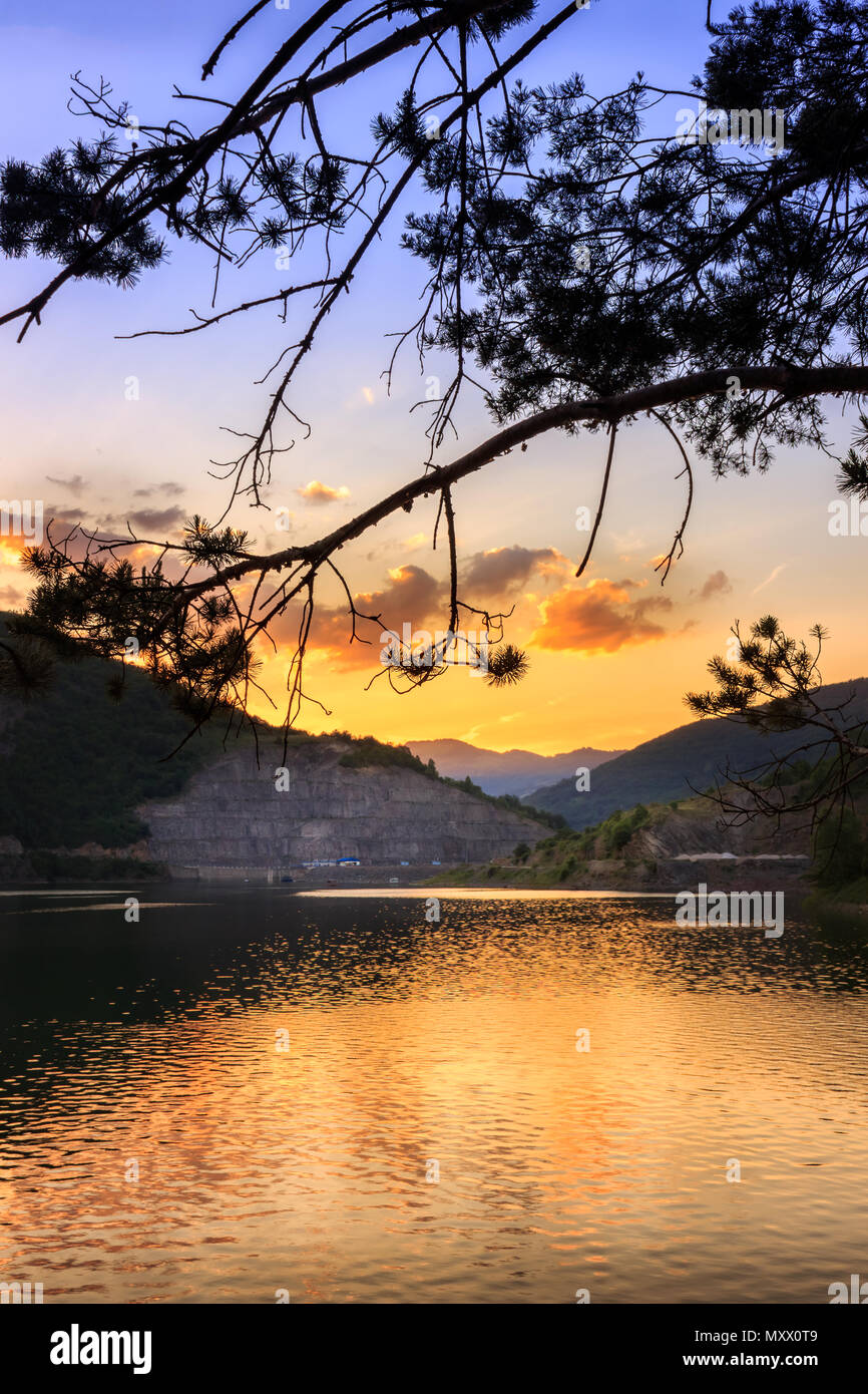 Pino silhouette, drammatica nuvole e cielo e brillante tramonto riflettente, setosa lago d acqua Zavoj, nei pressi di Pirot in Serbia Foto Stock