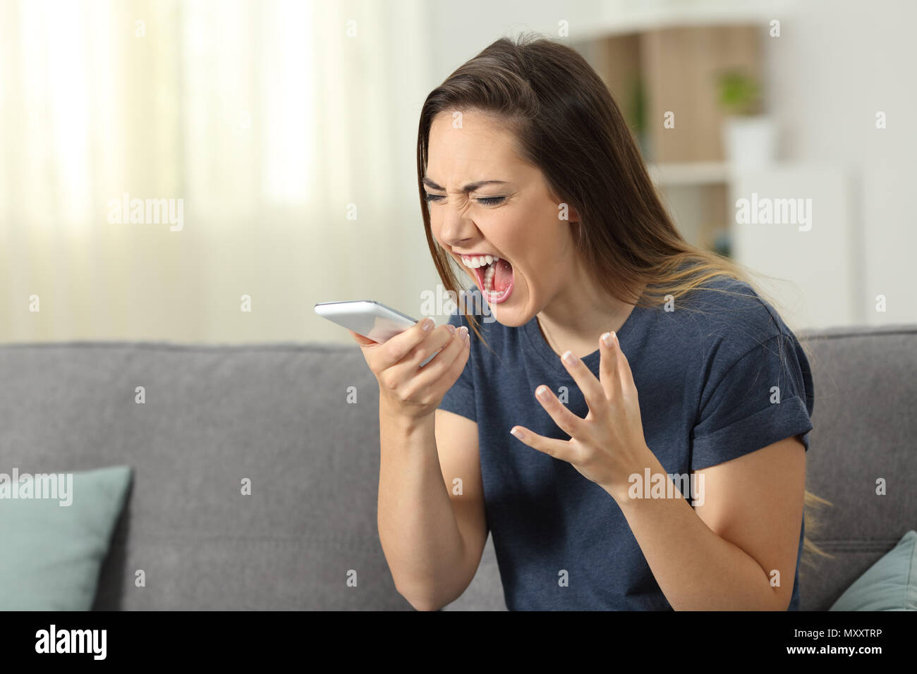 Donna furiosa gridando a smart phone seduta su un divano nel salotto di casa Foto Stock