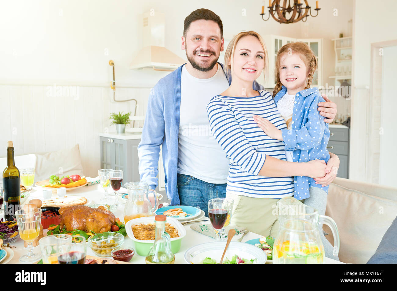 Ritratto di felice famiglia giovane con graziosi bambina in piedi alla cena di gala tabella con piatti deliziosi e sorridente alla fotocamera in moderno appartamento Foto Stock