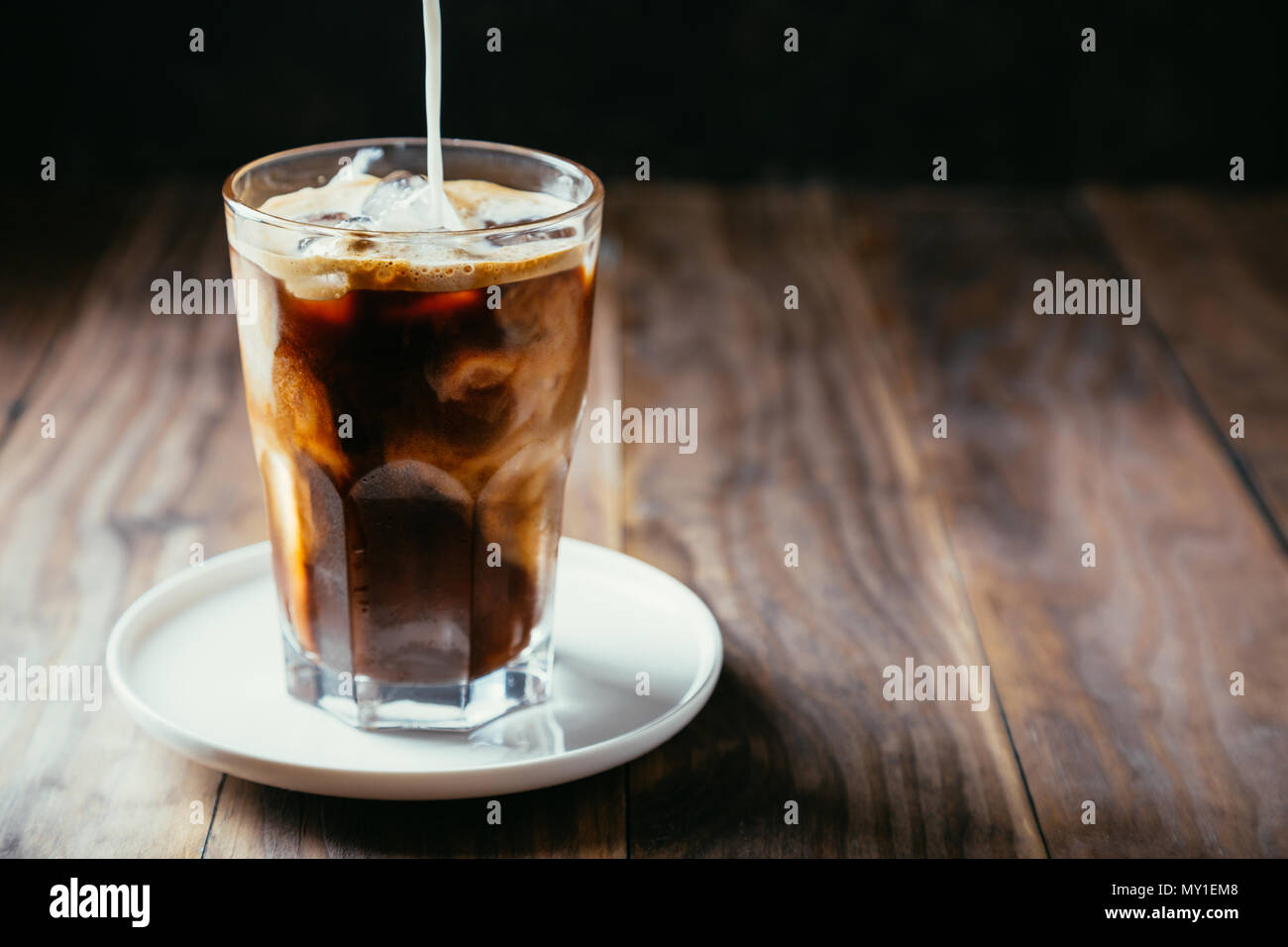 Versare il latte in un caffè ghiacciato in un bicchiere con una cannuccia su una tavola in legno rustico Foto Stock