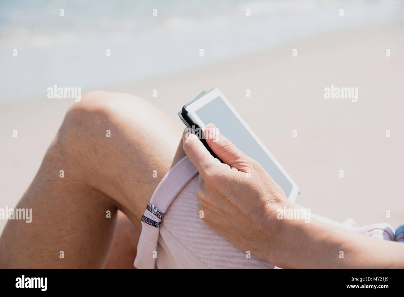 Primo piano di un giovane uomo caucasico sulla spiaggia, indossare pantaloncini rosa, utilizzando una tavoletta digitale Foto Stock