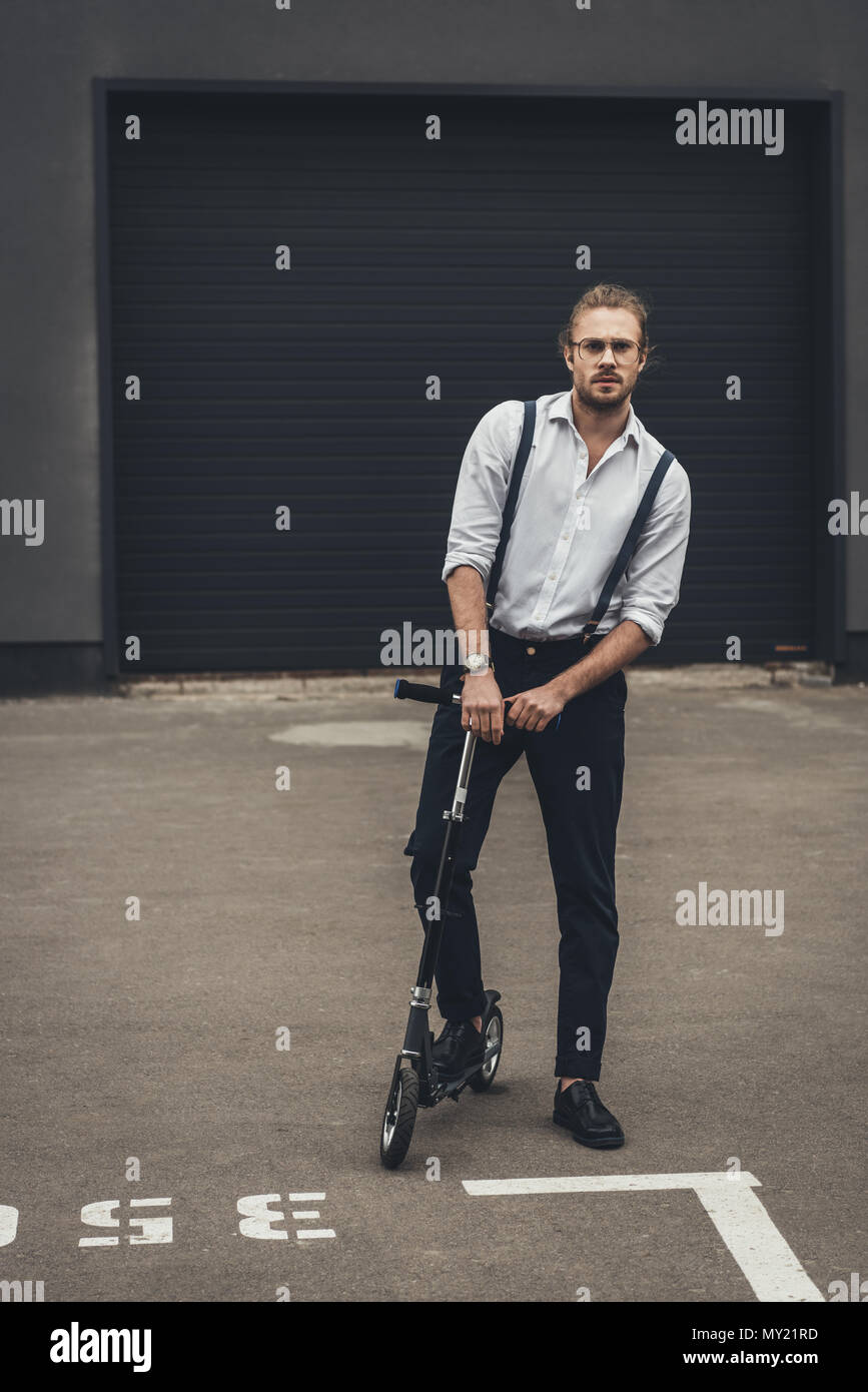 Elegante giovane uomo in occhiali e giarrettiere in piedi su scooter su parcheggio Foto Stock