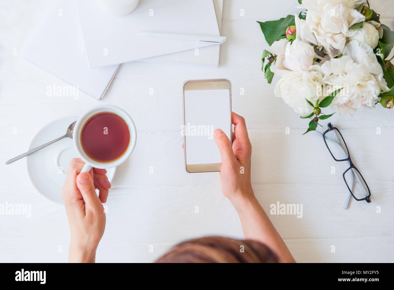Area di lavoro con la ragazza mani smart phone con schermo vuoto e la tazza di tè, bicchieri, notebook, peonia bianco sul tavolo ligneo. Vista superiore del femminile scrivania in ufficio. Pianificazione. Freelance di luogo di lavoro. Foto Stock
